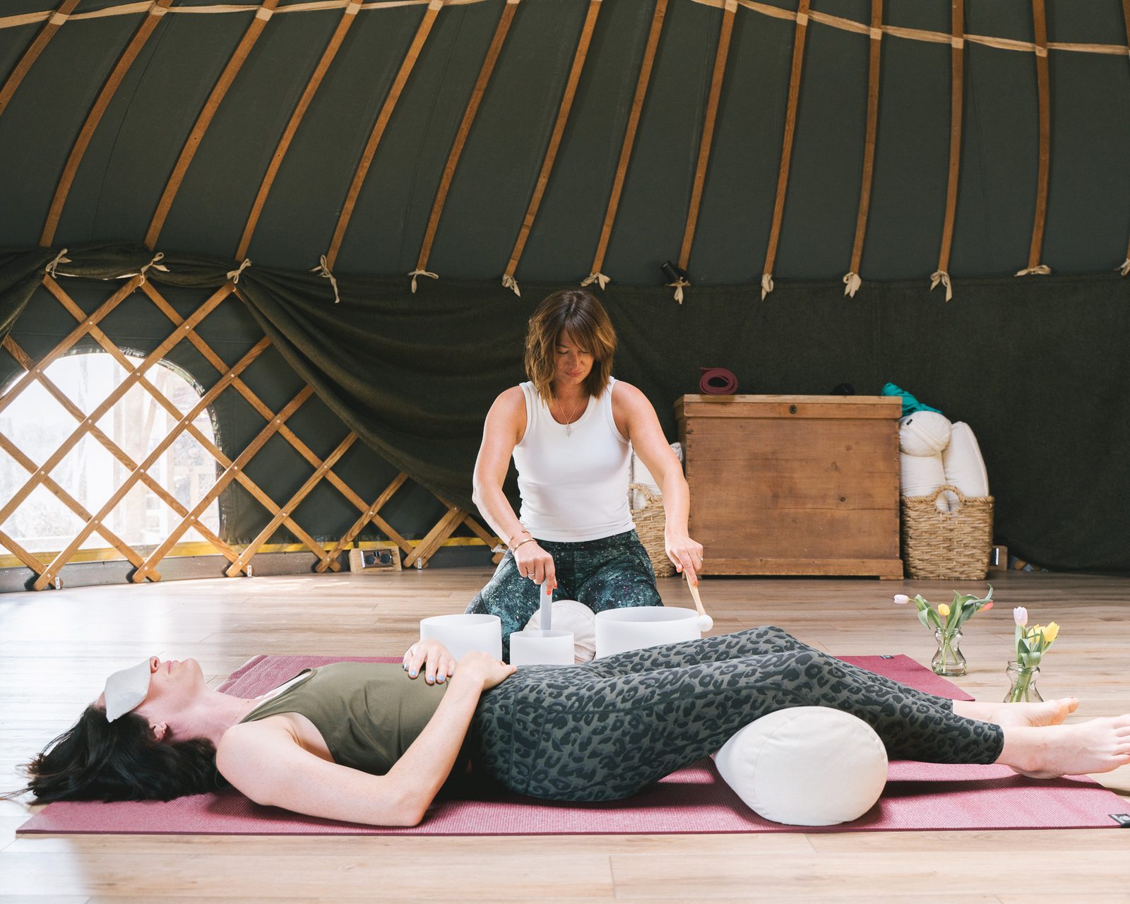 Lucy sounding a singing bowl over a student resting on a mat inside a yurt